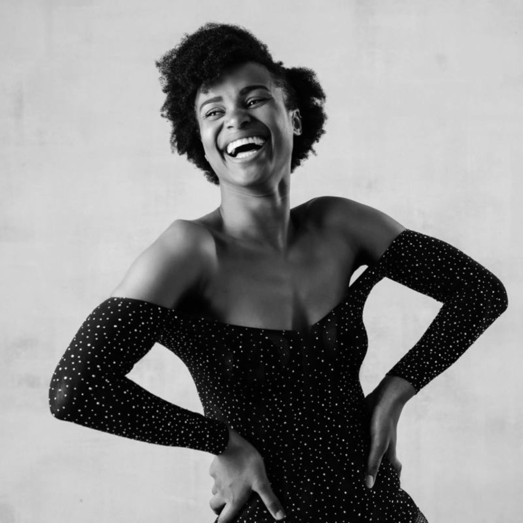 Pearl Cicci smiling softly at the camera, seated in a calm, minimalist space. She wears neutral tones and exudes a grounded, peaceful energy reflecting her work in somatic healing and nervous system support.